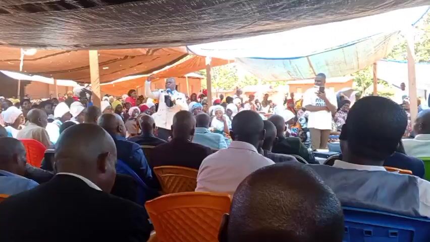 Certainly! Here's a description of the video:

The video shows a large group of people gathered outdoors under a makeshift canopy made of tarps. The majority of the crowd are seated on chairs, facing a man who is speaking into a microphone. He is wearing a light-colored suit. Many women in the audience have head coverings. A few people can be seen holding phones, presumably recording or taking pictures. The setting seems to be in a rural or semi-rural area, with simple structures visible in the background. The atmosphere appears to be attentive as the man delivers his message.