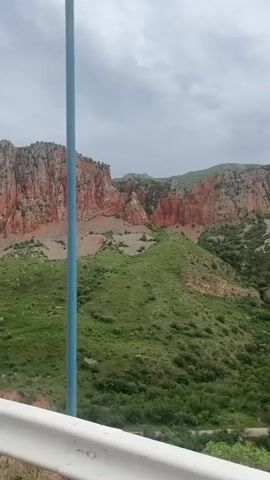 A camera pans left across a rocky mountain range with a narrow winding road visible at the bottom of the video frame. The mountain slopes are covered in green grass and shrubs. The rock face of the mountain has a reddish hue, with a significant amount of loose rock and scree at its base. The sky is overcast.