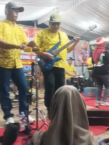 A video shows a band performing on a stage covered by a white tarp. The band consists of several men in matching yellow shirts with a black and red pattern. One man in the foreground is playing a blue electric guitar, while another to his left plays a small instrument that looks like a ukulele or a mandolin. Behind them, other band members are visible, including one playing drums and another on keyboard. On the stage, in front of the band, there are at least two women dancing. One woman in a light-colored dress is in the center, and another woman in a dark top and jeans is to her right, also dancing. There are also other people on the stage, including a man in a black shirt to the left of the dancers. The stage is covered in a red carpet, and in front of the stage, the audience is visible. The audience members are partially obscured, but one person in the foreground is wearing a beige hijab and a plaid shirt. The overall environment appears to be an outdoor event or a makeshift indoor venue. The lighting is somewhat dim, with overhead lights illuminating the stage. There are banners and posters visible in the background on the stage.