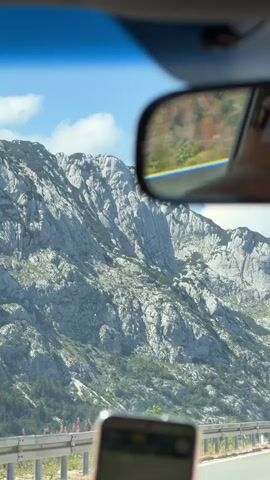 The video shows a view from inside a car. In the foreground, a smartphone is visible. In the middle ground, a rearview mirror reflects the back of a person's head wearing a brown hat with a logo, and a cross rosary hangs from the mirror. The background displays a mountainous landscape and a highway with a car driving in front.