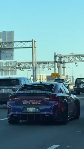 The video shows the rear of a blue and black Dodge Charger with custom bodywork, sitting in traffic on a highway. The car features graphics and lettering on the rear, including “SHE THICC”, a custom louvre, and license plate "SHETHIC".  The road is busy with other vehicles, including a Toyota SUV and a Jeep. Overhead road signs are visible. The sky is clear.