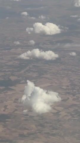 This is an aerial view of a landscape with scattered clouds. The ground appears to be agricultural land, with visible patterns of fields in varying shades of brown and tan. A few distinct clouds are visible, casting shadows on the ground below. The clouds are white and puffy, suggesting cumulus clouds. The perspective is from a high altitude, possibly from an airplane. No people or man-made structures are clearly discernible on the ground. The lighting suggests it is daytime.