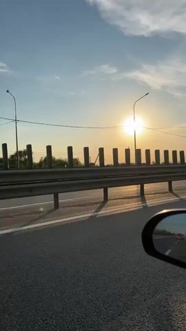 The video shows a side view from a car traveling on a highway during sunset. The sun is bright and low in the sky, casting a warm glow. A guardrail runs along the side of the road, with evenly spaced posts. Streetlights are visible on poles above the guardrail. In the rearview mirror, the reflection of the road and other cars can be seen. The sky is blue with scattered clouds. The road appears to be paved with asphalt, and there is a white line marking the lane. The overall impression is one of a peaceful drive as the day ends.