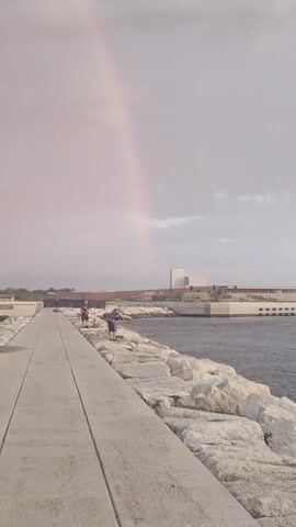 A vertical, wide shot of a partially formed rainbow over a body of water. In the foreground, a concrete walkway leads away from the camera, bordered by a rocky embankment on the right. Several people are visible on the walkway and rocky embankment, appearing to be fishing. In the background, there is a distant building with white walls and a long, brown structure. The sky is overcast with a faint pink hue.