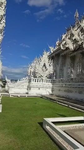 The video shows a panoramic view of the exterior of a white temple, likely a Buddhist temple, with intricate ornate designs and sculptures. The temple is set against a backdrop of a clear blue sky with scattered white clouds. In the foreground, there is a well-maintained green lawn and a paved walkway with a white border. The camera pans from left to right, revealing more of the temple's architecture, which features multiple tiered roofs, decorative eaves, and detailed carvings. The overall scene is serene and visually striking, highlighting the beauty and complexity of the temple's design.