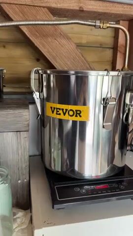 The video shows a distillation setup. There are several stainless steel pots, one with a "VEVOR" sticker on it, connected by flexible metal and plastic tubing. One pot sits on an induction cooktop, while another is elevated on a wooden stool. A large glass carboy filled with liquid is visible on the floor, and another smaller glass jar is on a lower shelf. The background consists of wooden paneling.