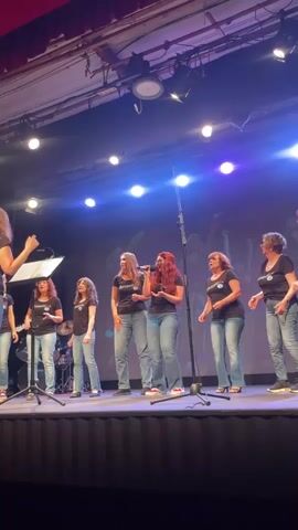 A group of seven women are standing on a stage, singing. They are all wearing black t-shirts and blue jeans. The woman in the center, with long red hair, is holding a microphone and singing. The other women are also singing, some with microphones and some without. There is a music stand in front of the woman on the far left, and a drum set in the background. The stage is lit with bright lights, and there is a dark curtain behind the singers. The video is shot from a low angle, looking up at the singers. The singers appear to be in a performance or rehearsal.