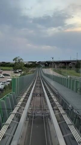 The video shows a first-person perspective of a monorail or automated people mover traveling along elevated tracks. The tracks are flanked by a green metal fence on the left and a similar fence and walkway on the right. Beyond the fence on the left, a parking lot with several cars and some trees can be seen. In the background, there is a highway overpass and distant buildings. The sky is cloudy, with some patches of light suggesting either sunrise or sunset. The lighting is dim, and the overall atmosphere is overcast. The motion is smooth, indicating the vehicle is moving at a steady speed.
