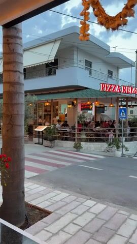 The video captures an early evening scene on a street in Ksamil, Albania, featuring a bustling corner restaurant and passersby. In the foreground, a decorative garland hangs above a tiled sidewalk, which leads to a zebra crossing with distinctive red and white stripes.

The prominent building on the left is a two-story structure, with the ground floor housing a restaurant named "PIZZA REI & RAI," indicated by a red neon sign. The restaurant has an inviting outdoor dining area, enclosed by a low fence, where several patrons are seated at tables, enjoying their meals. The upper floor of the building features balconies and white awnings.

Across the street, people are walking along the sidewalk and crossing the road. A white car drives past, and a white van is parked further down on the right. Another building with balconies and exterior lights is visible on the right side of the street. A tall utility pole with numerous wires stands near the center of the frame, and a "FARMACI" (pharmacy) sign is visible in the distance. The sky above is cloudy, suggesting a transition from day to evening. The overall atmosphere appears lively and relaxed.