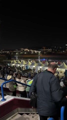 At night, a crowd of people is descending a staircase leading to an outdoor area filled with white tents. The area appears to be part of an event or festival, possibly a sporting event given the presence of a sports facility with lights in the background. Individuals are walking around, seemingly headed towards or away from the tents. Some people are standing on the stairs, observing the scene below. The lighting is mixed, with the tents illuminated from within and streetlights brightening the periphery.