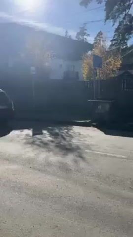 The video shows a street scene in a mountainous area on a sunny day. A black SUV drives away from the camera down a street lined with buildings and trees, towards snow-capped mountains in the distance. There are street signs and a pedestrian crossing sign visible on the side of the road. Two people are walking on the sidewalk in the opposite direction of the SUV. The road appears to be slightly worn, with visible cracks and patches.