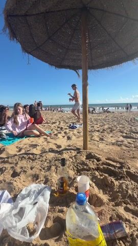 A sunny beach scene, viewed from underneath a thatched umbrella. There are many people scattered on the sand and in the ocean. A couple is standing at the shoreline. Near the camera are several empty bottles and a red bull can. People are sunbathing, walking along the beach, and playing in the water. The sky is blue with scattered clouds.