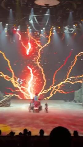 The scene is a live performance on a stage with a massive backdrop displaying dynamic red and white light effects, including streaks and lightning bolts. In the foreground, several performers are visible. One person is riding a motorcycle and executing a mid-air jump. Others are on motorcycles and an ATV adorned with stacked gift boxes. Several additional performers dressed in red costumes are also present on stage. The stage setup includes prop structures such as rocks and a gate. The audience, seen silhouetted at the bottom, is reacting with excitement. The lighting rig above the stage is visible, contributing to the overall ambiance of a theatrical show.