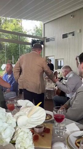 A man in a tan suit jacket stands with his back to the camera, facing a man playing a guitar at an outdoor gathering. The guitarist sits on a chair, playing his instrument. Several other people are seated at a table in the foreground, which is decorated with white flowers and food. The background shows a building with large windows and greenery outside.