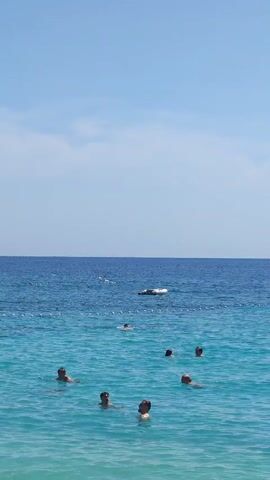The video shows a clear blue sky over a bright turquoise ocean. Several people are swimming in the water. In the distance, a white sailboat is anchored, and a small lighthouse stands on a rocky island. The water appears calm, with gentle waves. The scene suggests a sunny day at a beach or coastal area, with people enjoying recreational activities in the water.