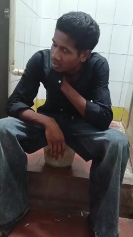 The video opens with a close-up shot of a young man with dark hair, wearing a black collared shirt and blue jeans. He is sitting on a low, circular object, possibly a bucket, and looking to his left with his hand resting on his chest. The background appears to be a tiled wall, possibly in a bathroom or similar utilitarian space, with a pipe visible on the left. The lighting is dim.

The camera then pans to the right, revealing another young man wearing a maroon collared shirt and black pants, crouching and looking at his phone. He is also seated on a similar low object. A portion of a black backpack is visible behind him. The floor is tiled with a reddish-brown hue and appears somewhat grimy, with some white debris scattered on it.

The view shifts again, showing the young man in the black shirt extending his arm towards the person in the maroon shirt. The person in the maroon shirt is looking down at his phone. The camera then zooms out slightly and shows the two young men interacting. The person in the black shirt's arm is extended, and their hands appear to be in the process of touching or shaking. The person in the maroon shirt is still looking at their phone. The background remains the same tiled wall and floor. The video concludes with the interaction between the two individuals.