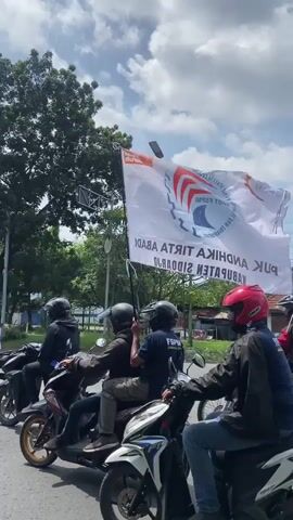The video shows a procession of motorcyclists on a city street. The motorcyclists are wearing helmets. Many are carrying white flags, some with blue symbols on them. A large white banner is also visible, with red and blue symbols and text. The riders are traveling under a partly cloudy sky, past buildings and trees. There are other vehicles and pedestrians visible. The overall scene suggests a parade or demonstration.