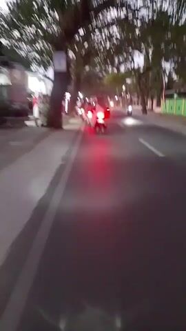 The video captures a perspective from a moving vehicle, likely a motorcycle or scooter, traveling on an asphalt road. The time appears to be dusk or early evening, as artificial lights are visible. Several other motorcycles, some with passengers, are also on the road, all moving in the same direction. Their taillights are illuminated, casting red glows on the road. On the left side of the road, there is a sidewalk, lined with trees and some structures that resemble shops or stalls. Some of these structures have lights on. The overall scene suggests a busy street in a town or city during the evening commute or a social gathering. The video is shaky, indicating the motion of the camera.