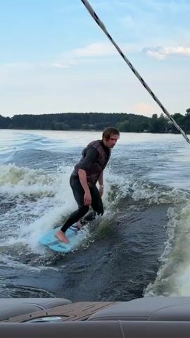 A person in a wetsuit and life vest is wakesurfing behind a boat. They are standing on a light blue wakesurf board and riding the wake of the boat. The water is choppy, and the sky is cloudy with some blue visible. Trees line the distant shore. The camera appears to be positioned on the back of the boat, looking towards the wakesurfer.
