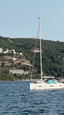 A white sailboat with a tall mast sails on blue water. The boat has a light blue canopy over its cockpit where several people are visible. In the background, a green, tree-covered hill slopes down to the water. Several white buildings are scattered across the hillside. The sky is clear and blue.