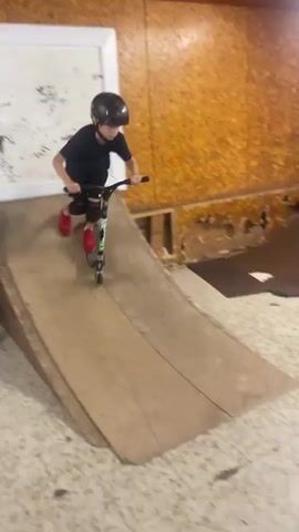 A young person wearing a dark blue t-shirt, black shorts, red shoes, a helmet, and knee pads is riding a scooter in a skate park. The skate park has wooden ramps and structures. The person rides the scooter towards a ramp and then up and over it, performing a trick. The video is captured from a slightly elevated angle.