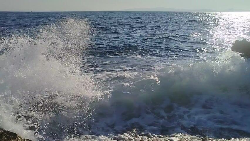 This media file depicts a dynamic ocean scene with waves crashing against rocks. The camera appears to be positioned at an unusual angle, possibly underwater or looking up from the water, as the scene is upside down. The subjects are the natural elements of the sea: the deep blue water, the white foam of the breaking waves, and dark, rugged rocks.

The activity is the powerful movement of the ocean, with waves in various stages of formation and breaking. The water is turbulent, creating spray and froth as it interacts with the rocky shore. Sunlight glints off the surface of the water and catches the mist, creating a sparkling effect.

Notable details include the clear distinction between the dark, solid rocks and the fluid, energetic water. The weather appears to be bright, with strong sunlight indicating a clear or partly cloudy day. The location is clearly coastal, with the presence of rocks and the sea. The overall mood is one of natural power and the raw beauty of the ocean. There is no visible text in the media file. The scene suggests a dramatic moment of wave action at the coast.