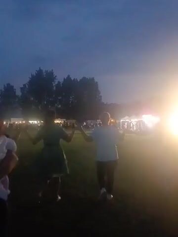 Three adults, two women and one man, hold hands and walk in a circle on a grassy field in the foreground. Another partially visible person on the far left holds a baby. In the background, an outdoor event with multiple lit tents and numerous people is visible under a dark evening sky. Trees are present in the mid-ground, and a bright light source emanates from the right side of the background.