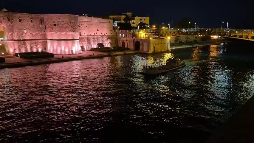 A nighttime aerial shot shows a boat navigating a body of water, with a castle illuminated by pink lights on the left side of the screen. The castle walls are made of stone, with turrets and ramparts visible. The water is dark, with reflections of the lights from the castle and surrounding buildings shimmering on its surface. The boat is small, with a few people on board. The overall atmosphere is serene and picturesque.