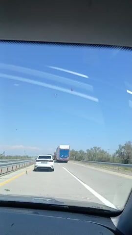 The video shows a view from inside a car looking out the windshield at a highway. The sky is bright blue and clear. There are several vehicles on the road, including a white car in front of the camera car, and multiple large trucks. One truck appears to be a car carrier, with the structure for holding vehicles visible. The truck in front of the car carrier is a semi-trailer truck. The vehicles are traveling in the same direction. There are trees and vegetation along the side of the highway, and a guardrail is visible. The sun appears to be high in the sky, suggesting it is daytime. The overall scene is one of normal highway traffic on a clear day.