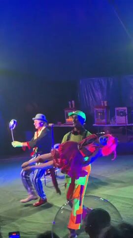 A dimly lit circus tent is illuminated by vibrant neon blue and purple lights. In the center of the scene, a performer dressed in a brightly colored clown costume with a hat is walking from left to right, carrying a person dressed as a doll. The doll-like figure has bright pink hair and a ruffled outfit. Another performer, wearing a green hat and a neon green shirt with orange and neon green striped pants, stands to the right of the clown, holding the doll-like figure in their arms. The background shows a dark tent ceiling with some lighting equipment visible. There are also some illuminated booths or stands with popcorn machines in the distance. The audience is partially visible in the foreground, with some people holding up their phones to record the performance. The overall atmosphere appears to be a circus show with a focus on neon and glow-in-the-dark elements.