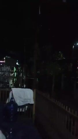 The video shows a nighttime scene, possibly outdoors. A bright light, possibly a streetlamp or floodlight, illuminates a dark area with trees and what appears to be a structure in the background. Some faint, colored lights are also visible, possibly decorative. The foreground shows a wooden fence and a white cloth draped over something. The video is shaky and the image is somewhat blurred, suggesting it was filmed handheld in low light conditions.