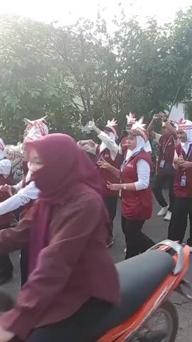 A group of young people, dressed in maroon vests over white shirts and black pants, and wearing white headbands with red and white decorations, are performing a dance in the street. Some of them are also wearing white hijabs. They appear to be participating in some kind of parade or festival. The background shows trees and a street. A motorcycle is parked on the left side of the frame. The lighting suggests it is daytime, possibly morning or late afternoon, with sunlight filtering through the trees. The individuals are engaged in synchronized movements, raising their hands and gesturing, indicating a choreographed performance. Some individuals have identification badges clipped to their vests. The overall atmosphere seems festive and energetic.