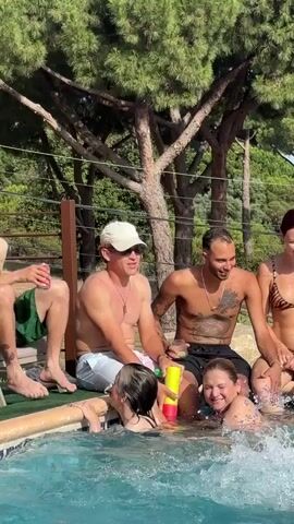 Six adults, three male and three female, are gathered around a swimming pool on a sunny day. Three men are sitting on the edge of the pool, one holding a can, one wearing a white hat, and another with tattoos on his chest and arms. Three women are in the pool, two interacting with a man holding a water gun. The background features lush green trees and a wooden fence. The scene depicts a casual social gathering by a pool.