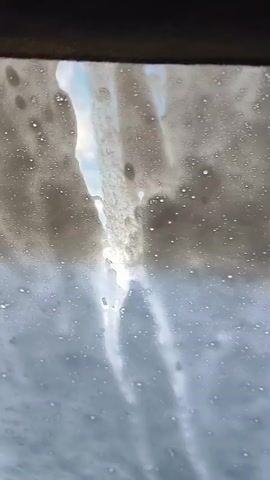 The video shows a close-up view of water and soap suds running down a glass surface. The suds are white and foamy, and the water is clear. The glass appears to be a window or windshield, and the context suggests it is being washed. The light filtering through the suds creates a bright, hazy effect.