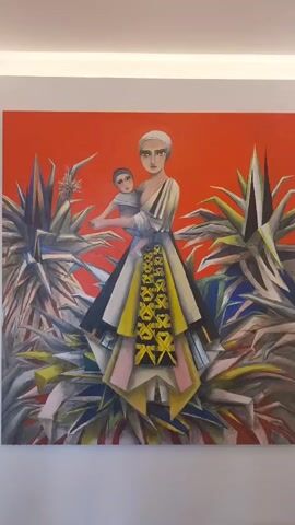 This media file shows a painting of a woman holding a baby. The figures are rendered in a stylized, almost abstract manner, with sharp geometric lines and a limited color palette. The woman wears a head covering and a layered dress with a prominent yellow and black patterned panel. She is surrounded by large, spiky plants, which also have a geometric quality. The background is a solid, vibrant orange. The overall impression is one of strength and perhaps a sense of being in a foreign or exotic landscape. The style of the painting suggests it is modern art. There are no visible text elements in the media file.