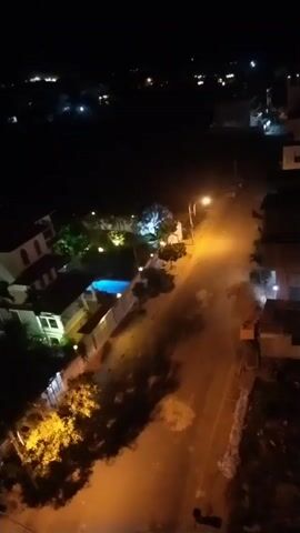 A high-angle, slightly shaky video shows a residential street at night. The street is lit by a streetlamp. On the left side of the street are several houses, one with a swimming pool. The houses are illuminated by exterior lights. Trees are visible in front of the houses. On the right side of the street, there are some unlit structures, possibly under construction. In the distance, lights from other buildings can be seen in the darkness.