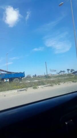 The video, filmed from the inside of a moving car, shows an exterior daytime scene. The sky is blue with scattered white clouds. The landscape features a flat, sandy area with sparse vegetation and some buildings in the distance. Power lines run overhead. A white pickup truck is seen driving on a road parallel to the car. A child is sitting in the bed of the truck. In the background, there is smoke rising into the air. The car's dashboard is visible in the foreground.