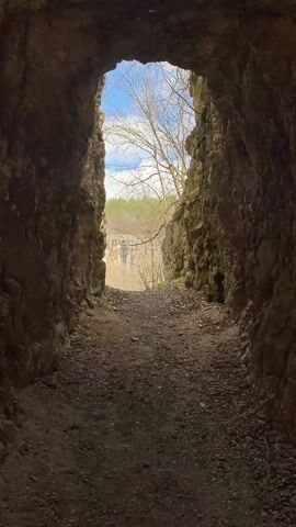 The camera looks down a tunnel cut through rock and debris. The tunnel slopes slightly uphill and the path is covered with pebbles and dirt. Outside of the tunnel is a cliff with some trees and blue sky with white clouds.