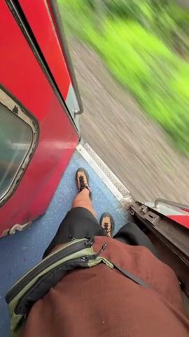 The video begins with a first-person perspective shot from inside a train, looking out the open door. A person's legs, wearing black shorts and tan sandals, are visible standing on the train's blue floor. A brown shirt and a green and black fanny pack are also visible. The train is red and appears to be moving, as indicated by the blurred green scenery and tracks outside. The camera then pans to the right, showing a large, blurry pile of reddish-brown material, possibly construction debris or waste, against a cloudy sky. Green grass is visible in the foreground, also blurred due to motion. The video ends with a quick pan across the debris pile.