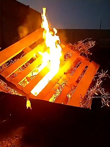 The video shows a fire burning on top of a wooden pallet in an outdoor setting. The pallet is angled upward, and the flames are visible in the spaces between the wooden slats. There is some dry brush underneath the pallet. In the background, there is a building and a dark sky. An indiscernible noise, possibly speech, is present.