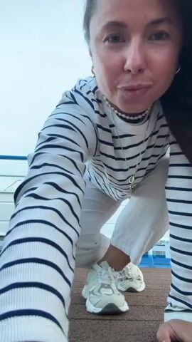 A woman with long, dark hair wearing a striped long-sleeved shirt and white pants is dancing on a deck. She is wearing white sneakers. The deck appears to be on a boat, with a white railing in the background. The sky is cloudy and blue. The woman is moving her arms and legs in a fluid motion, her hair blowing in the wind. She is smiling at the camera. The video captures her in various poses as she dances.