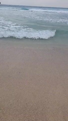 The video shows a beach with a sandy shore and waves gently rolling in. The water is a light blue-green color, and there are whitecaps on the waves. The sand is a tan color. The sky is overcast.