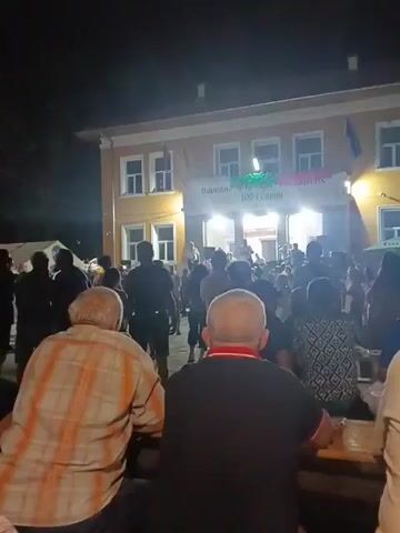 The video opens with a wide shot of a nighttime outdoor gathering in front of a light-colored, two-story building. The building appears to be a community center or school, with flags and a banner displayed across its facade. The banner has text and possibly imagery, but it's difficult to read clearly from this distance and lighting. A crowd of people is gathered in front of the building, some standing and some seated at tables. The atmosphere seems festive, with lights illuminating the scene and suggesting an event or celebration is taking place. The camera focuses on the crowd from behind, showing their backs as they look towards the building. In the foreground, two older men are seated at a table. The overall impression is one of a community event happening after dark.