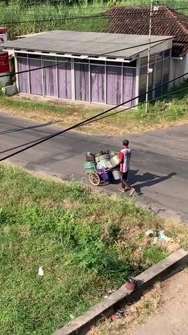 A person is pushing a cart loaded with various items, including containers and what appears to be metal objects, along a paved road. The person is walking away from the camera. To the left of the road, there is a building with a red and white sign. Further down the road, a motorcycle with two riders is approaching. The area is lined with green vegetation. Overhead, black cables are strung across the scene. The day is sunny, casting shadows on the road.
