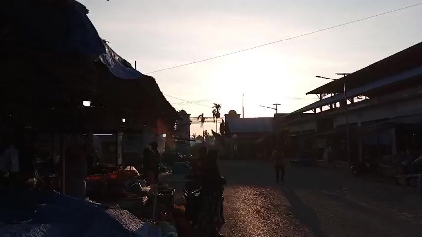 The video shows a street scene at a market during sunset. The sun is low in the sky, casting long shadows. On the left side of the street, there are market stalls with blue and white tarps, displaying various goods, some of which appear to be food items. Several people are visible near these stalls, some appearing to be vendors and others shoppers. On the right side of the street, there is a building with a sign that reads "PASAR IKAN," indicating a fish market. This building has a covered walkway with more stalls. Several motorcycles are parked or being ridden on the street. The ground is paved with cobblestones. The overall atmosphere suggests a bustling local market as the day draws to a close.