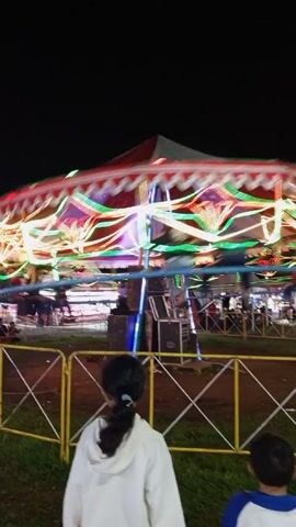 At night, in what appears to be a carnival or fairground setting, the camera captures a vibrant carousel in motion. The carousel is adorned with a red and white striped top, and is illuminated with numerous colorful lights that create streaks as it spins. In the foreground, two children are observing the ride. One is a girl with her hair pulled back, wearing a white hooded jacket, and the other is a young boy in a blue and white shirt. A simple yellow and white fence separates the children from the carousel. Sound equipment, including speakers and boxes, are situated near the carousel’s center. The ground consists of grass and dirt. Other people and carnival structures are faintly visible in the background.