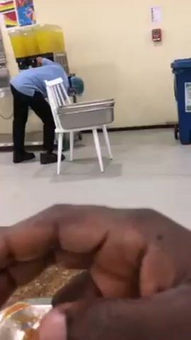 Inside a cafeteria-style dining area, a diner is eating a partially consumed meal from a metal tray. In the background, a person wearing a blue shirt, black pants, and a surgical cap or hairnet is working near a beverage dispenser. They are using a white chair to access something under the dispenser. A stainless steel container rests on the chair. A fire alarm is visible on the wall.