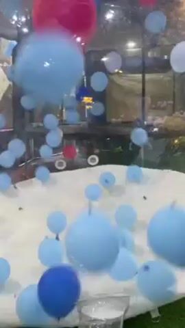 Here is a description of the video:

The video shows a clear balloon filled with many smaller balloons of various colors including light blue, dark blue, white, yellow, and red. The camera is inside the clear balloon, moving around to capture the scene. Outside the balloon, one can see a building decorated for a celebration, and several indistinct figures are present. The surrounding area is lit, suggesting an indoor location. A song plays in the background.