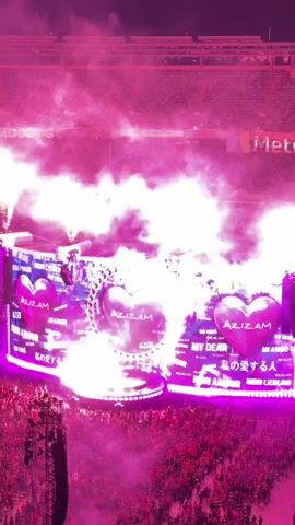 A concert stage is illuminated with pink and purple lights, with a large circular screen at the center displaying a heart-shaped image. The screen is surrounded by smaller heart-shaped images and text that reads "AZIZI," "MERA PYAAR," and "MY DEAR." Smoke and confetti fill the air above the stage, and a crowd of people can be seen in the foreground. The scene appears to be taking place at night, with stadium seating visible in the background.