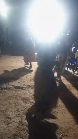The video shows a dimly lit outdoor area, possibly at night, with several people present. The scene is illuminated by bright overhead lights, creating stark shadows. The ground appears to be dirt. The people are dressed in various clothing, including what looks like long robes or dresses, and some are wearing masks or face coverings. They appear to be moving or dancing to music playing in the background. There are chairs visible in the background, suggesting an event or gathering is taking place. The overall atmosphere is somewhat mysterious due to the darkness and masking.